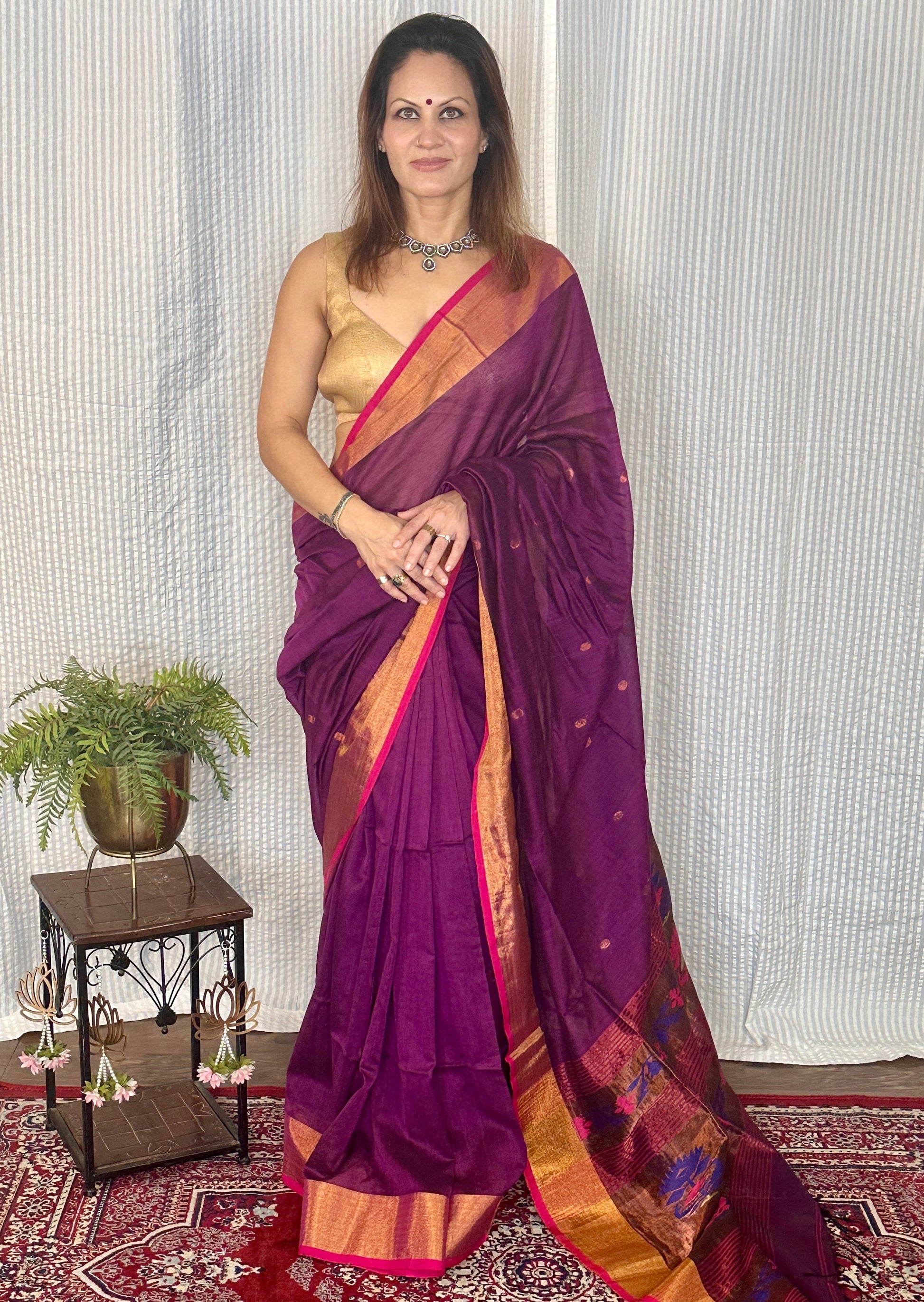 Woman wearing a purple saree with gold border indoors on a patterned rug.
